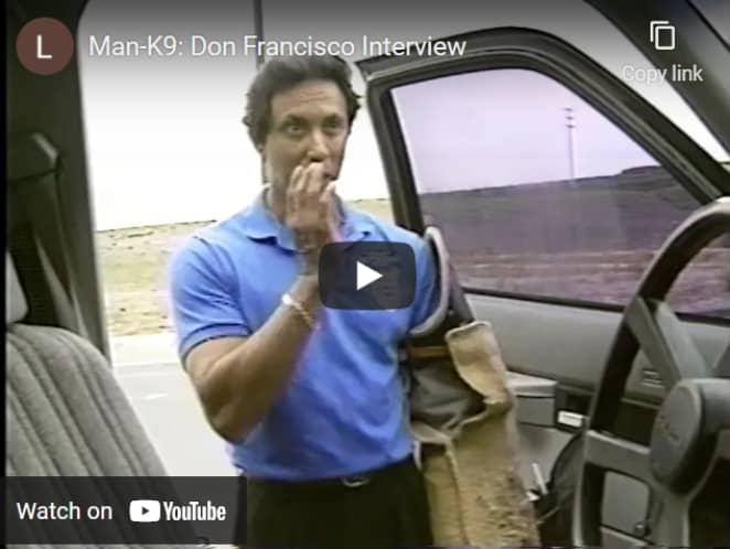 A man in a blue shirt standing by an open vehicle door gestures toward his face during an outdoor interview.