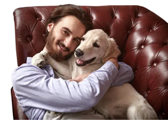A man with a beard and a light blue shirt cuddles a smiling golden retriever while sitting on a red leather sofa.