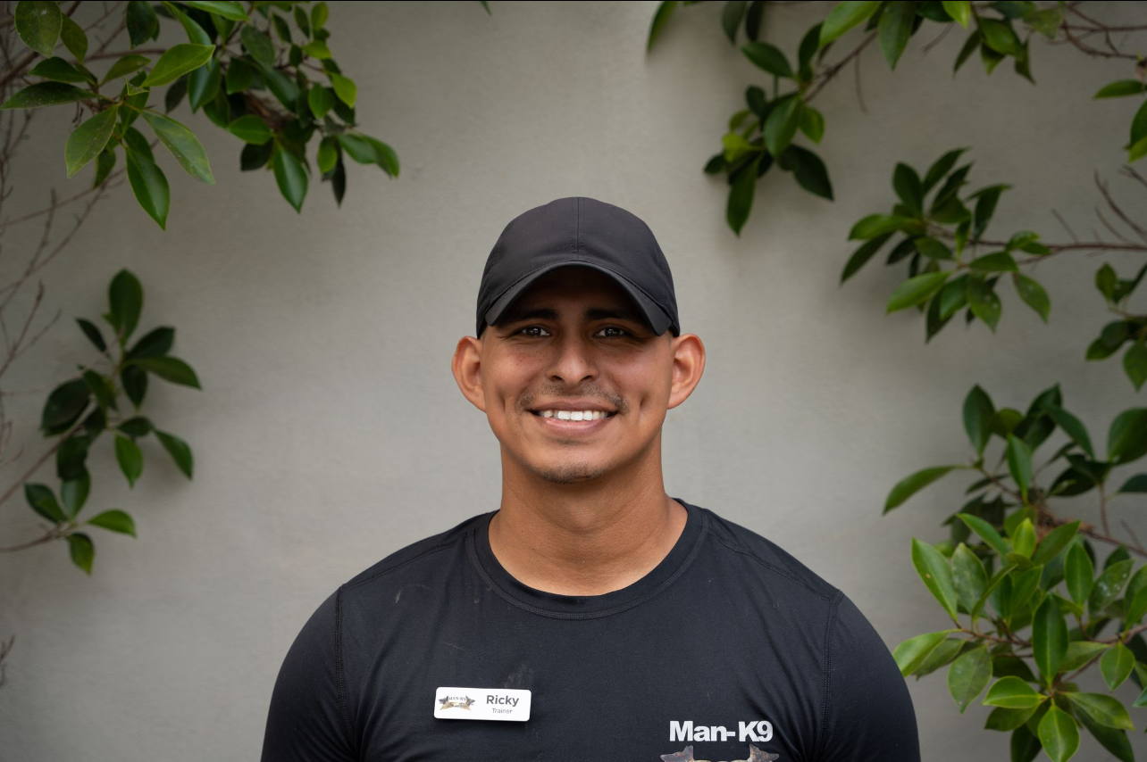 A person wearing a black shirt and cap smiles at the camera. The person is standing against a light-colored wall with green leaves in the background. The shirt has text reading "Man-K9.